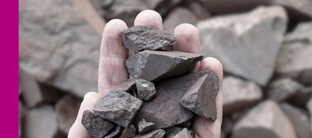 A hand holding a large handful of dark, reddish-brown, angular chunks of raw material, likely iron ore, bauxite, or mineral rocks.