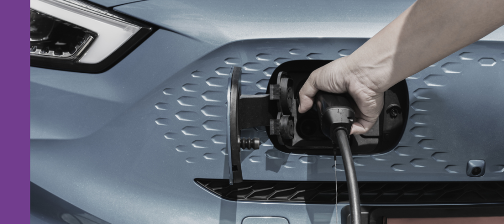 A person's hand plugging a black charging cable connector into the charging port located on the front grille area of a light blue electric vehicle.