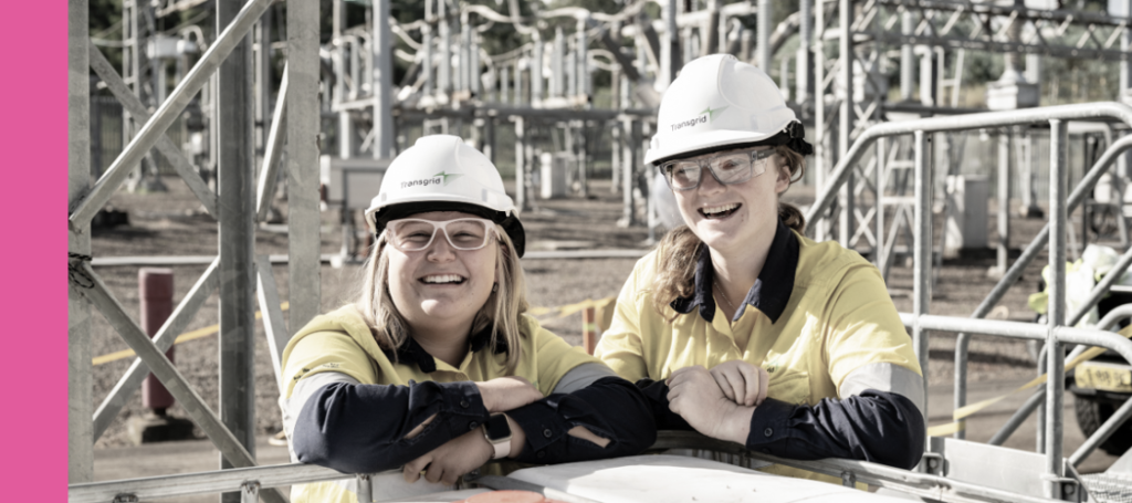 Two young women, both smiling and looking at the camera, posing at an electrical substation.